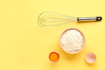 Bowl with wheat flour, cracked egg and whisk on yellow background