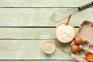 Composition with bowl of wheat flour, sesame seeds, eggs and whisk on green wooden table