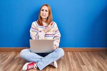 Young woman using laptop at home sitting on the floor pointing aside worried and nervous with...