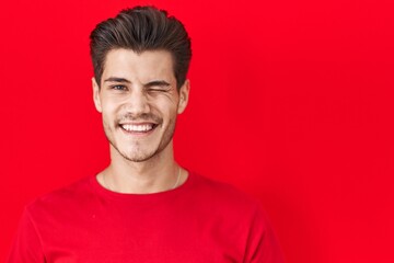 Young hispanic man standing over red background winking looking at the camera with sexy expression,...