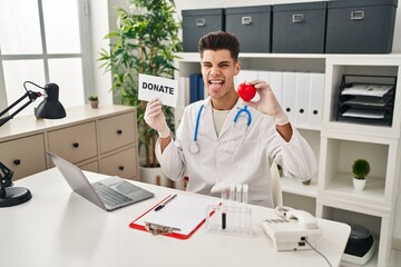 Young hispanic doctor man supporting organs donations sticking tongue out happy with funny expression.