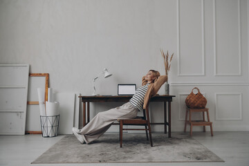 Relaxed young woman holding hands behind head while sitting at her creative working place