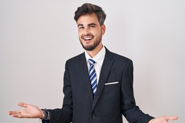 Young hispanic man with tattoos wearing business suit and tie smiling cheerful with open arms as friendly welcome, positive and confident greetings