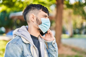 Young hispanic man wearing medical mask talking on smartphone at park