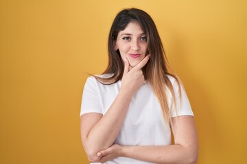 Obraz premium Young brunette woman standing over yellow background looking confident at the camera with smile with crossed arms and hand raised on chin. thinking positive.