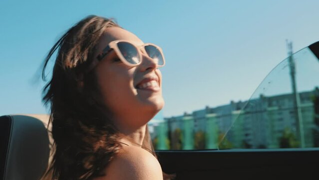 Portrait of young beautiful and smiling hipster female in convertible car. Sexy carefree woman driving cabriolet. Positive model riding and having fun in sunglasses at sunset