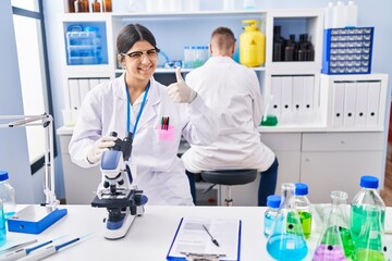 Young woman working at scientist laboratory smiling happy and positive, thumb up doing excellent and approval sign
