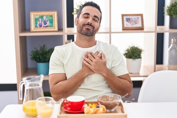 Hispanic man with beard eating breakfast smiling with hands on chest, eyes closed with grateful gesture on face. health concept.