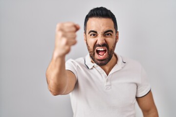 Young hispanic man with beard wearing casual clothes over white background angry and mad raising fist frustrated and furious while shouting with anger. rage and aggressive concept.