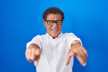 Middle age hispanic man standing over blue background pointing to you and the camera with fingers, smiling positive and cheerful
