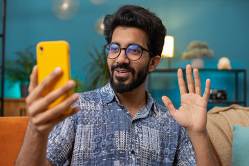 Happy indian man blogger taking selfie on smartphone, communicating video call online with social media subscribers followers, recording vlog stories. Young hindu guy at home apartment room on couch