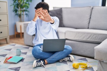 Non binary person studying using computer laptop sitting on the floor covering eyes with hands and doing stop gesture with sad and fear expression. embarrassed and negative concept.