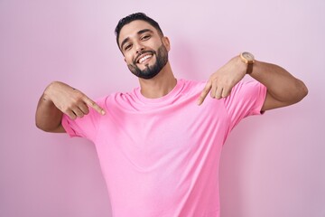 Hispanic young man standing over pink background looking confident with smile on face, pointing...