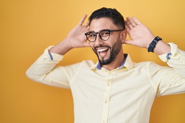 Hispanic young man wearing business clothes and glasses smiling cheerful playing peek a boo with hands showing face. surprised and exited