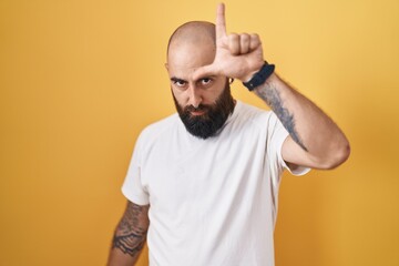 Young hispanic man with beard and tattoos standing over yellow background making fun of people with...