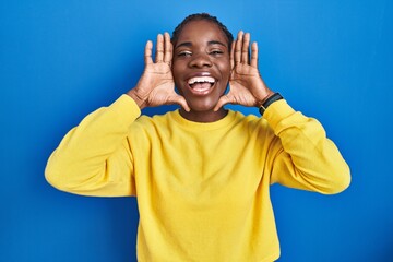 Beautiful black woman standing over blue background smiling cheerful playing peek a boo with hands...