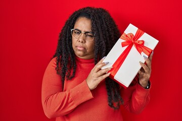 Plus size hispanic woman holding gift skeptic and nervous, frowning upset because of problem. negative person.