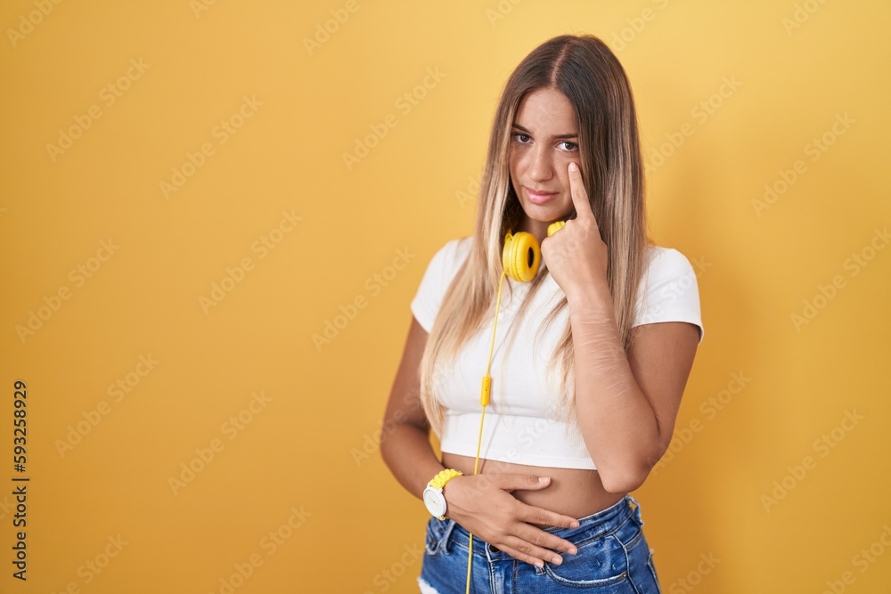 Poster young blonde woman standing over yellow background wearing headphones pointing to the eye watching y