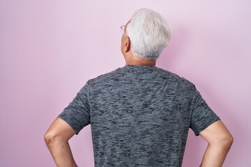 Middle age man with grey hair standing over pink background standing backwards looking away with...