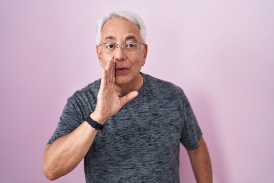 Middle age man with grey hair standing over pink background hand on mouth telling secret rumor, whispering malicious talk conversation