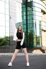 Fototapeta na wymiar Full length image of a young female model with long hair, in black sport clothes and white shirt, dance moves outside. 