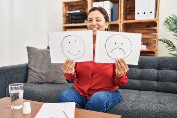 Middle age hispanic woman working on depression holding sad to happy emotion paper winking looking...