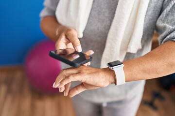 Middle age hispanic woman wearing sportswear using smartwatch and smartphone at laundry room