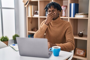 Young african man with dreadlocks working using computer laptop smelling something stinky and disgusting, intolerable smell, holding breath with fingers on nose. bad smell