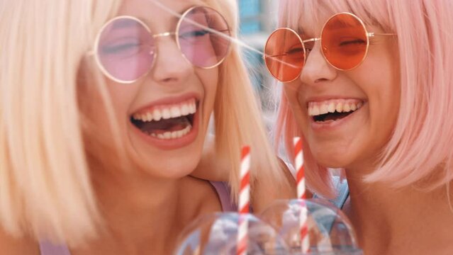 Two young beautiful smiling hipster female in trendy summer clothes. Sexy carefree women posing in the street in wigs. Positive funny models drinking fresh cocktail smoothie drink in plastic cup 