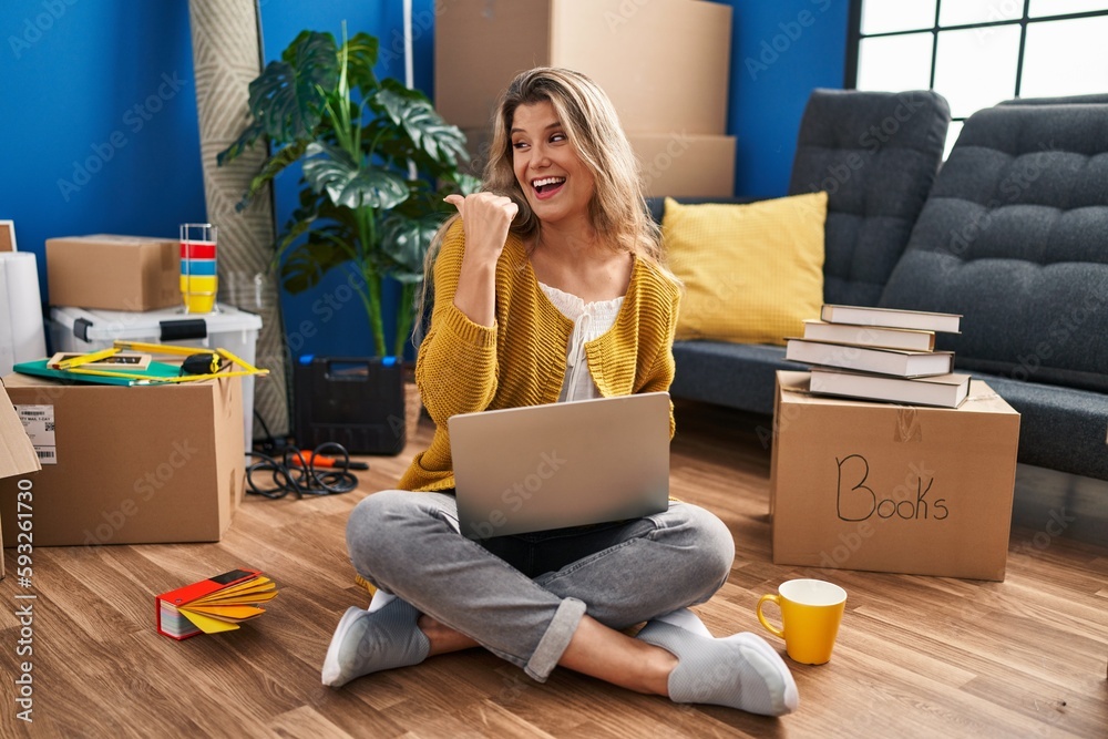 Sticker young woman sitting on the floor at new home using laptop smiling with happy face looking and pointi