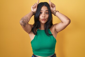 Brunette woman standing over yellow background doing funny gesture with finger over head as bull...