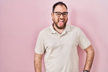 Plus size hispanic man with beard standing over pink background sticking tongue out happy with...