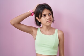 Naklejka premium Young girl standing over pink background confuse and wondering about question. uncertain with doubt, thinking with hand on head. pensive concept.