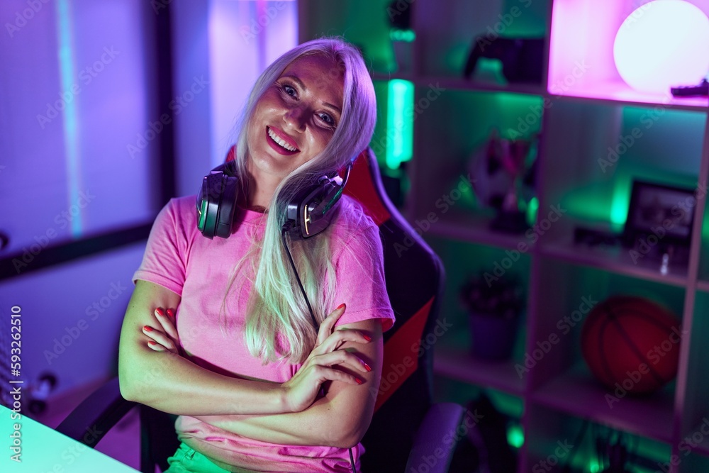 Sticker Young blonde woman streamer wearing headphones sitting with arms crossed gesture at gaming room