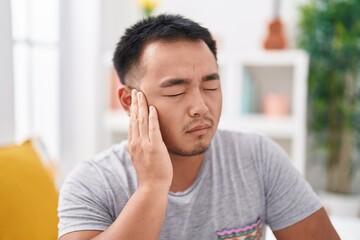 Young chinese man suffering for ear pain sitting on bed at bedroom