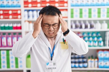 Chinese young man working at pharmacy drugstore suffering from headache desperate and stressed because pain and migraine. hands on head.