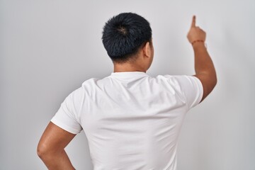Young chinese man standing over white background posing backwards pointing ahead with finger hand