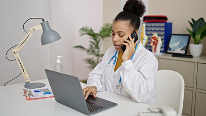 Young african american woman doctor using laptop talking on telephone at clinic