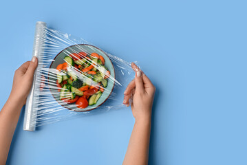 Female hands wrapping bowl with salad by stretch wrap on blue background