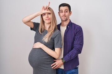 Young couple expecting a baby standing over white background making fun of people with fingers on...