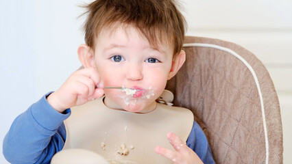 A happy child eats porridge with a spoon while sitting on a high chair. Baby in a bib eats oatmeal...