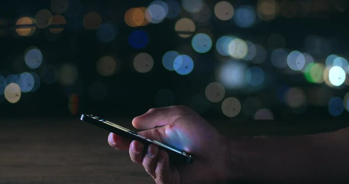 Male hand scrolling browsing social networks on phone on background of lights of night city. Cinematic shot male scroll through news feed on mobile app on smart phone. Scrolling social media on phone