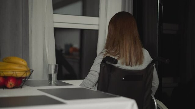 Woman with paralysis in wheelchair moves to kitchen window. Young housewife rests at home looking at street on weekend evening calmly backside view