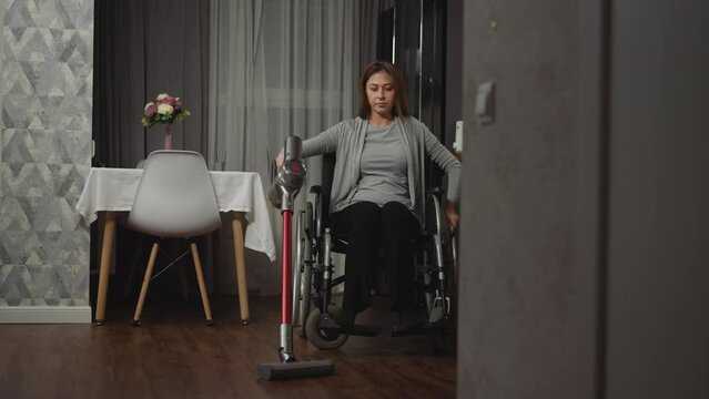 Woman with paralysis removes dust in house using convenient device. Young woman with injury uses medical equipment to move while doing household chores