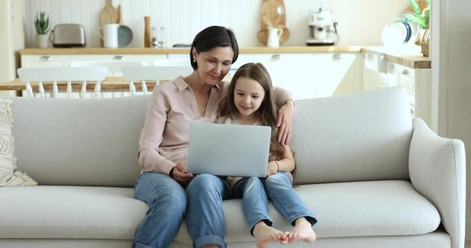 Cheerful mature grandmother and gen Z grandchild girl using laptop together, sitting close on couch at home, watching funny movie, making video call, looking at display, laughing