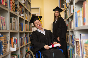 Happy young woman and woman in wheelchair in graduate gown with diploma in hands in library....