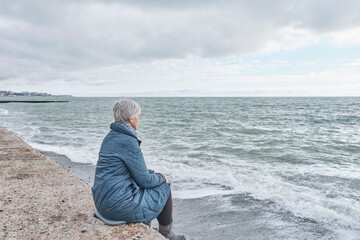 Senior woman sitting on sea coast on cold cloudy day.