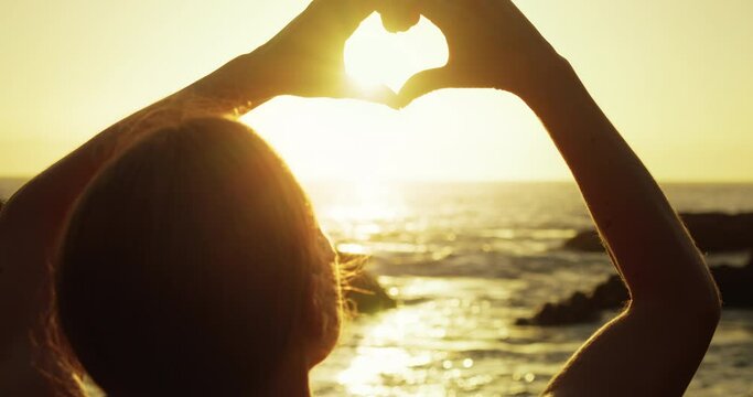 Girl on beach makes heart symbol with hands at sunset light. Summer vacation travelers dream. Sunlight glow between fingers.