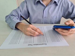 Closeup of teenager sitting at desk holding phone in her hand and taking exam test