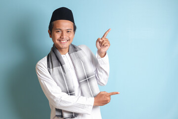 Portrait of young Asian muslim man showing product and pointing with his hand and finger to the side. Isolated image on blue background